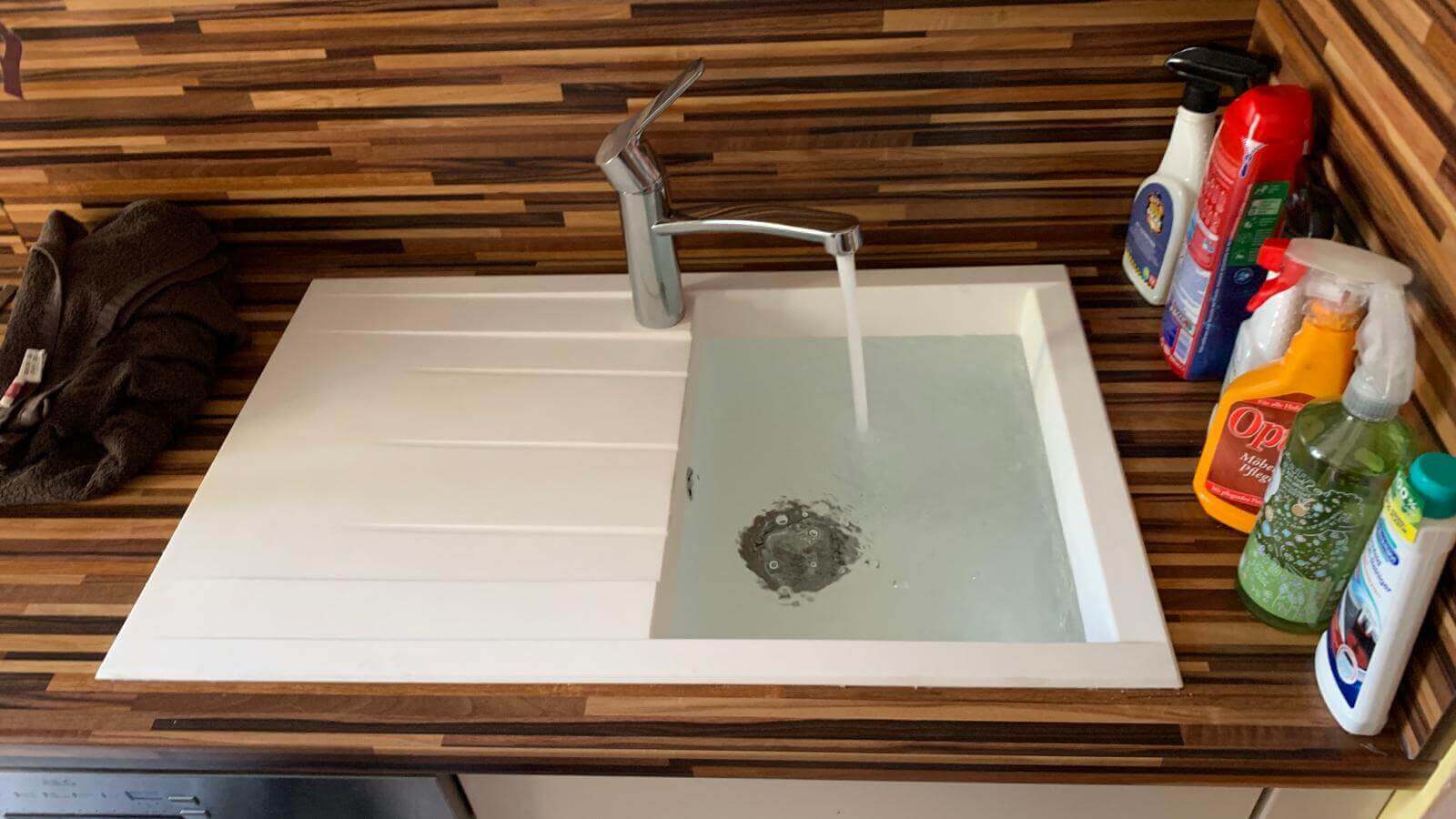 Moderne Küchenspüle mit laufendem Wasser, umgeben von Reinigungsmitteln auf einer Holzarbeitsplatte.