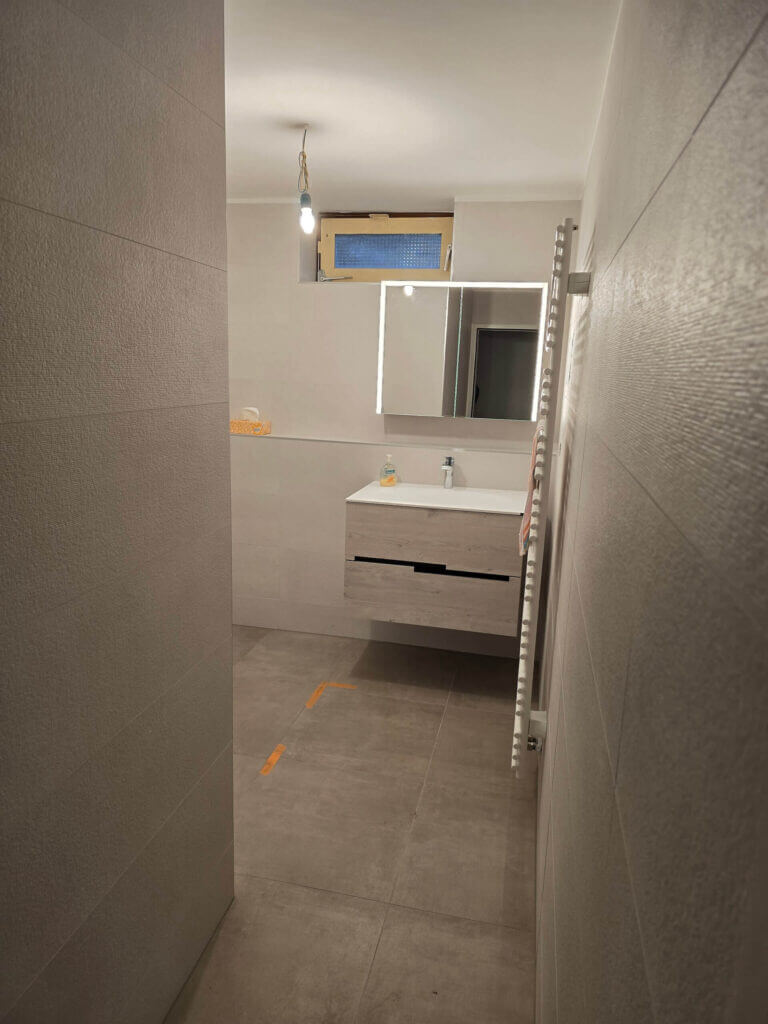 Modernes Badezimmer mit grauen Wandfliesen, beleuchtetem Spiegel und Hängelampe über Waschbecken.