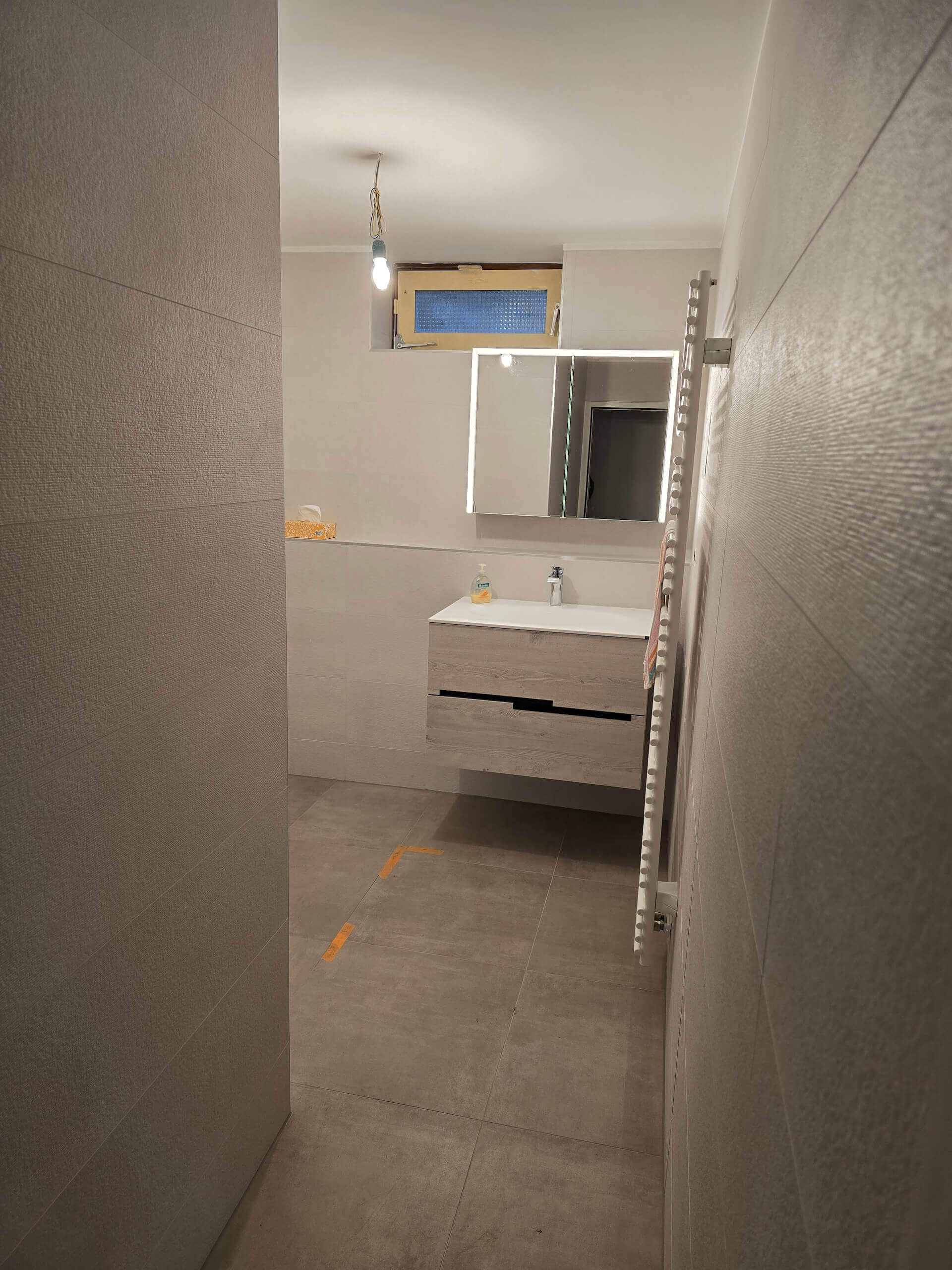 Modernes Badezimmer mit grauen Wandfliesen, beleuchtetem Spiegel und Hängelampe über Waschbecken.