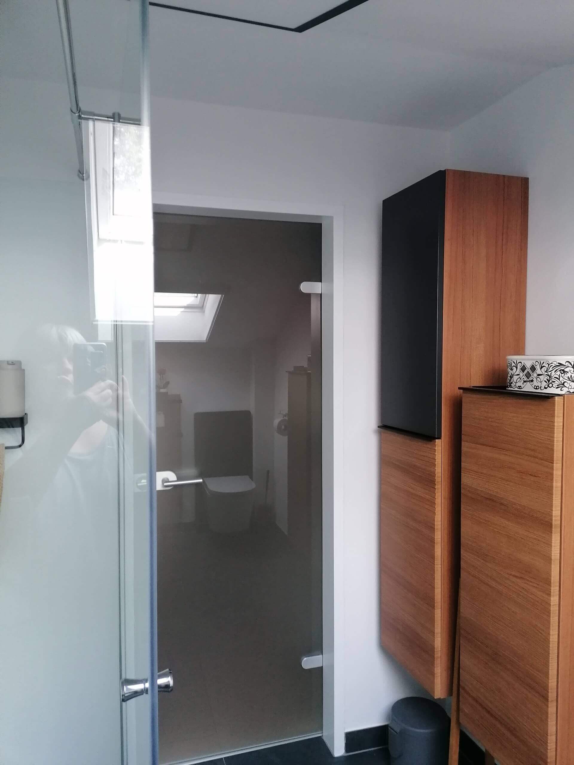 Modernes Badezimmer mit Holzschränken, Glastür und Dachfenster für natürliche Beleuchtung.