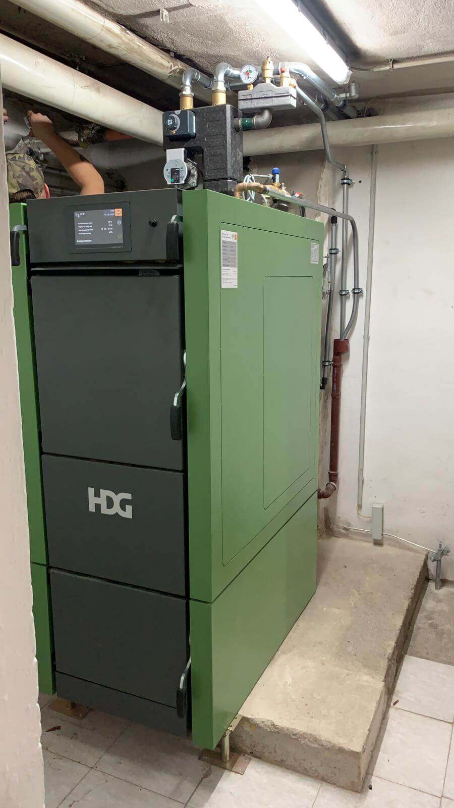 Grüne Heizanlage in einem Kellerraum, Wasserleitungen und ein Techniker im Hintergrund für Sanitärinstallationen.