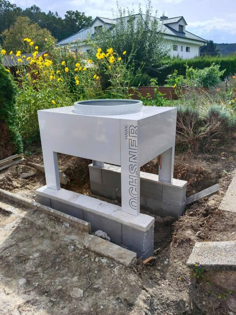 Moderne Heizungsinstallation im Garten vor Einfamilienhaus, umgeben von blühenden Pflanzen.