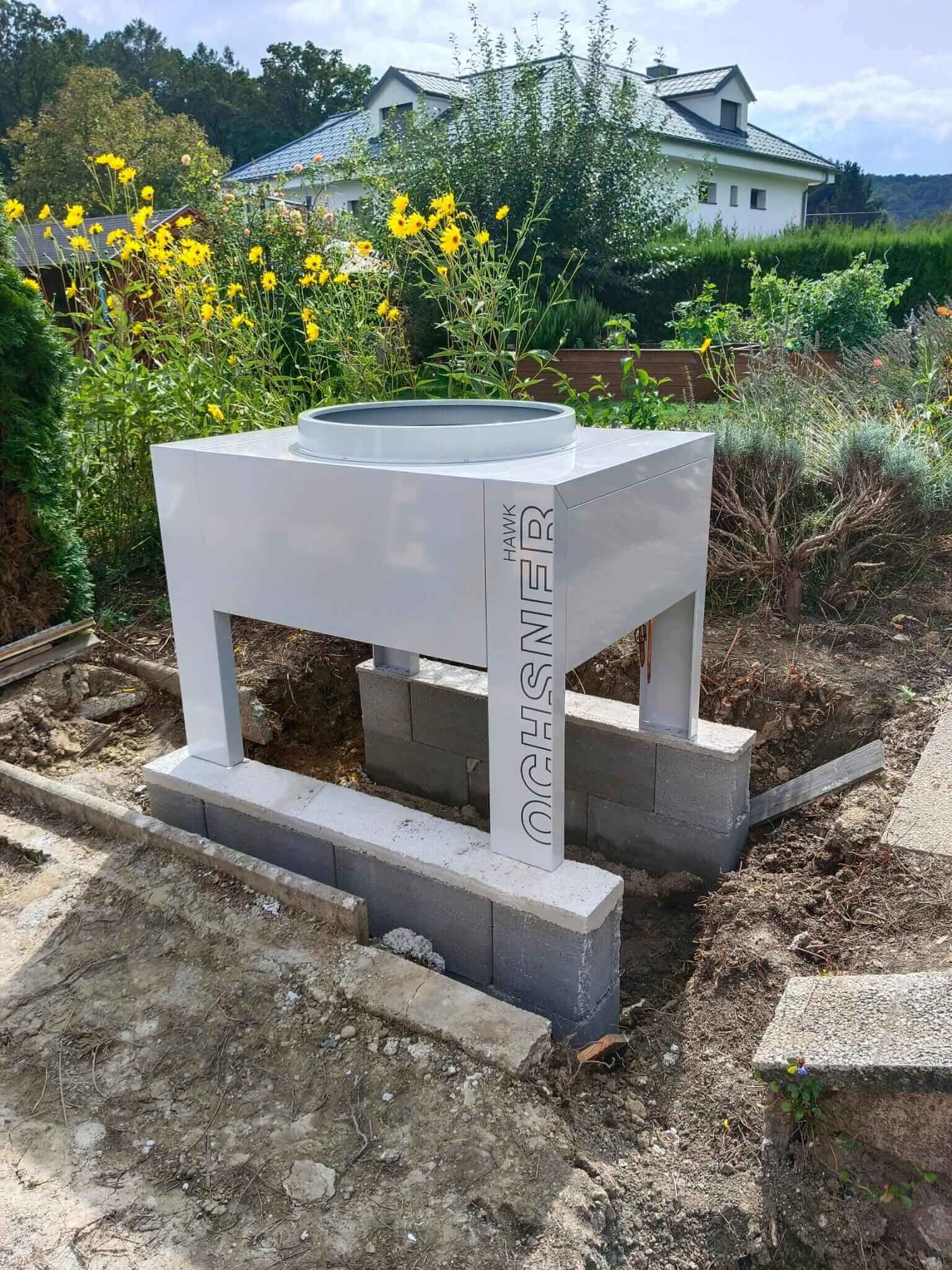 Moderne Heizungsinstallation im Garten vor Einfamilienhaus, umgeben von blühenden Pflanzen.
